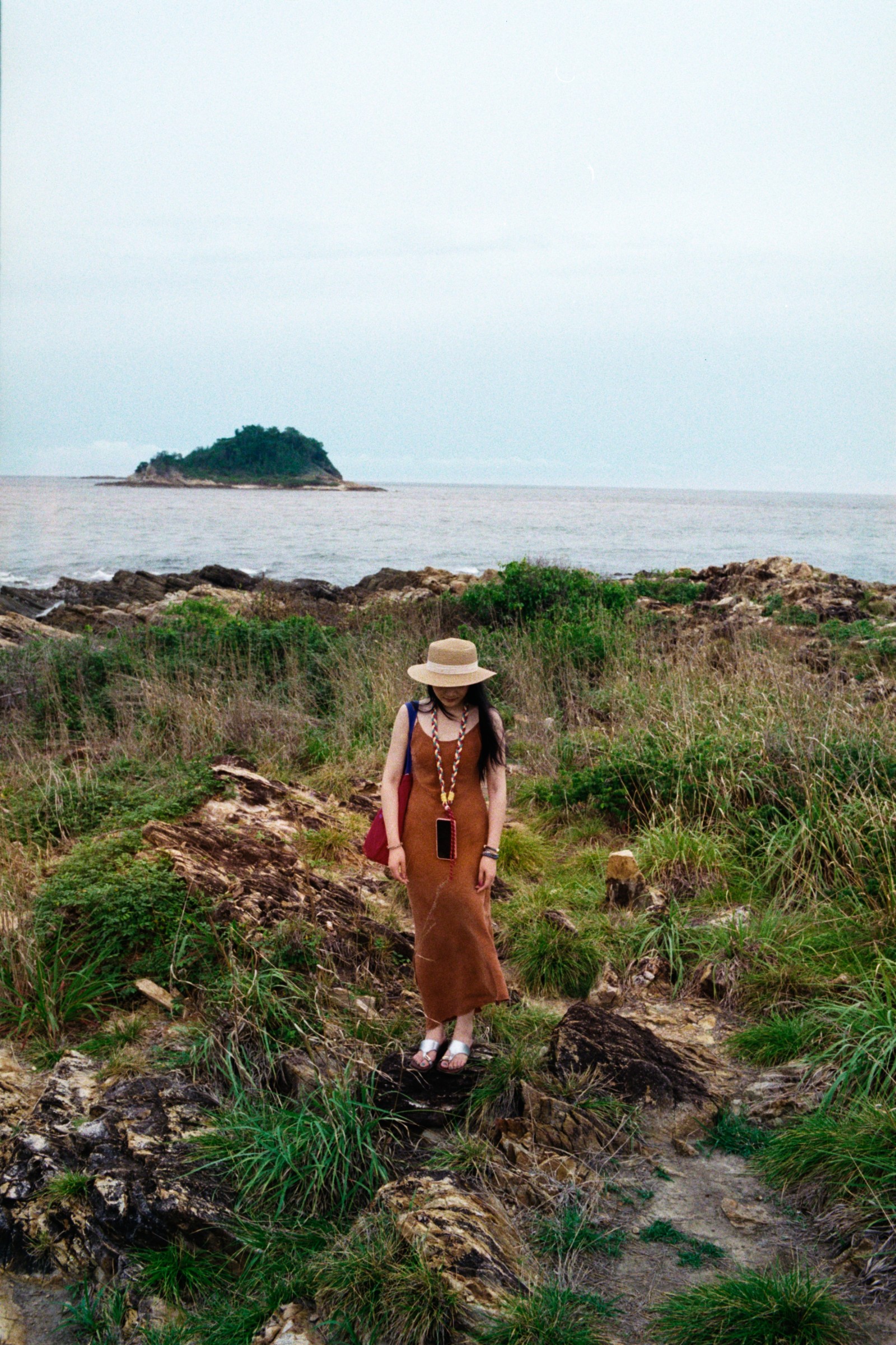 Woman in a brown dress and sunhat walking along a rocky coast